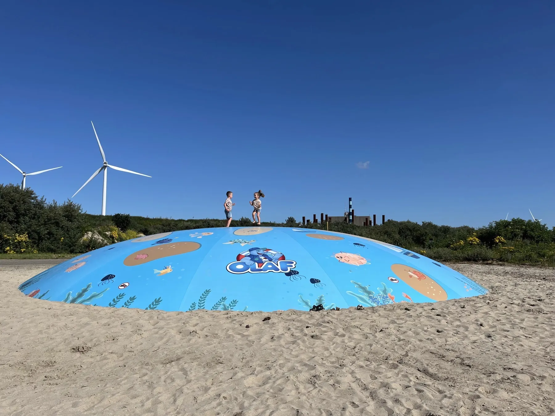 Twee kinderen springen hand in hand op een luchtkussen bij Deltapark Neeltje Jans met een windmolen op de achtergrond.