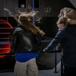 Bezoekers staan in de stormsimulator bij Deltapark Neeltje Jans en ervaren windkracht 12 met wapperende kleding en lachende gezichten.