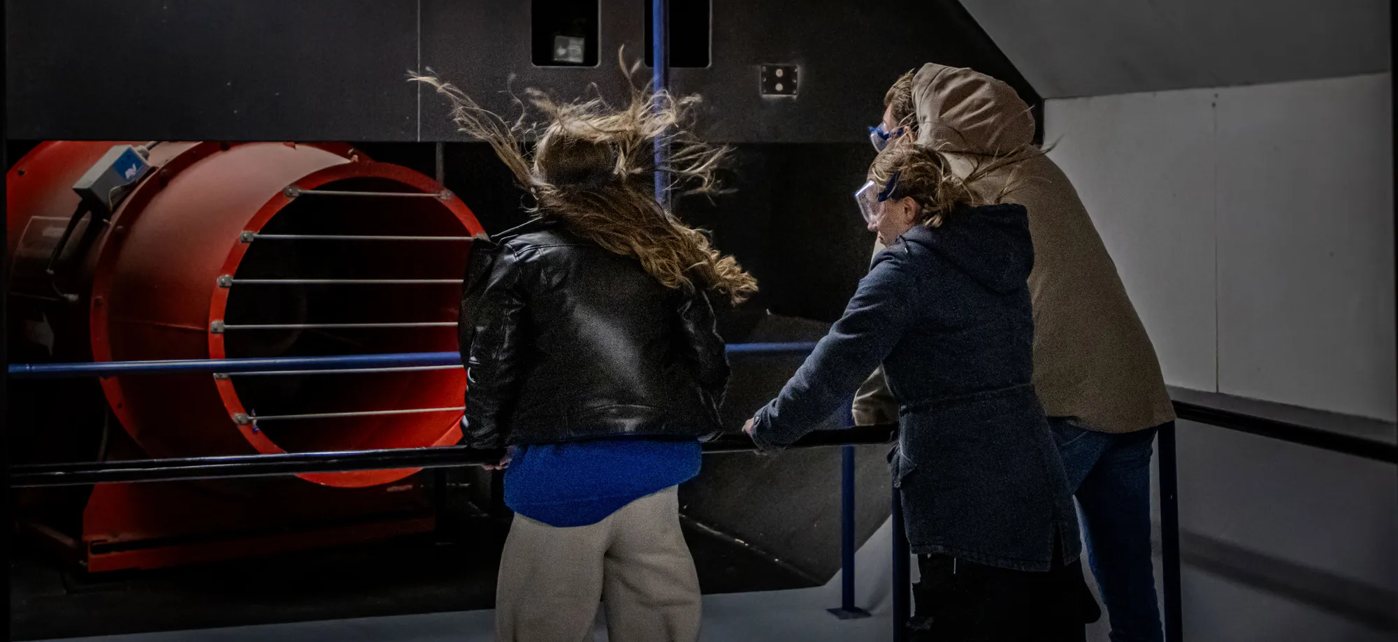 Bezoekers staan in de stormsimulator bij Deltapark Neeltje Jans en ervaren windkracht 12 met wapperende kleding en lachende gezichten.