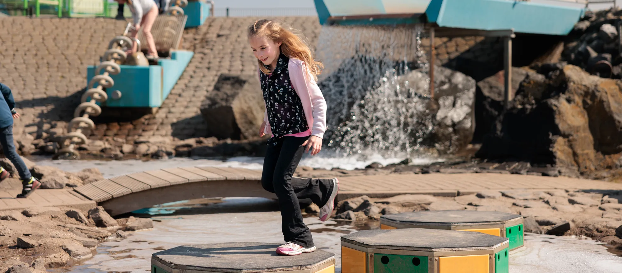 Lachend meisje rent over houten stapstenen in een interactieve waterspeeltuin bij Deltapark Neeltje Jans.