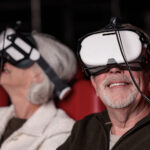 Twee oudere bezoekers met VR-brillen genieten van een virtuele ervaring in een theaterzaal met rode stoelen.