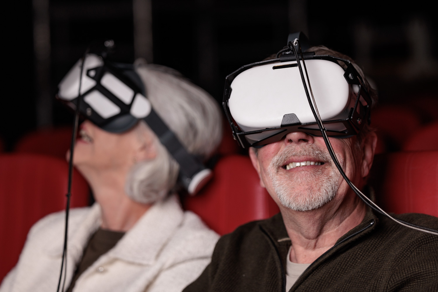 Twee oudere bezoekers met VR-brillen genieten van een virtuele ervaring in een theaterzaal met rode stoelen.