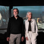 Oudere man en vrouw staan in een tentoonstelling over de Watersnoodramp van 1953, met projectie van een overstroomd dorp op de achtergrond.