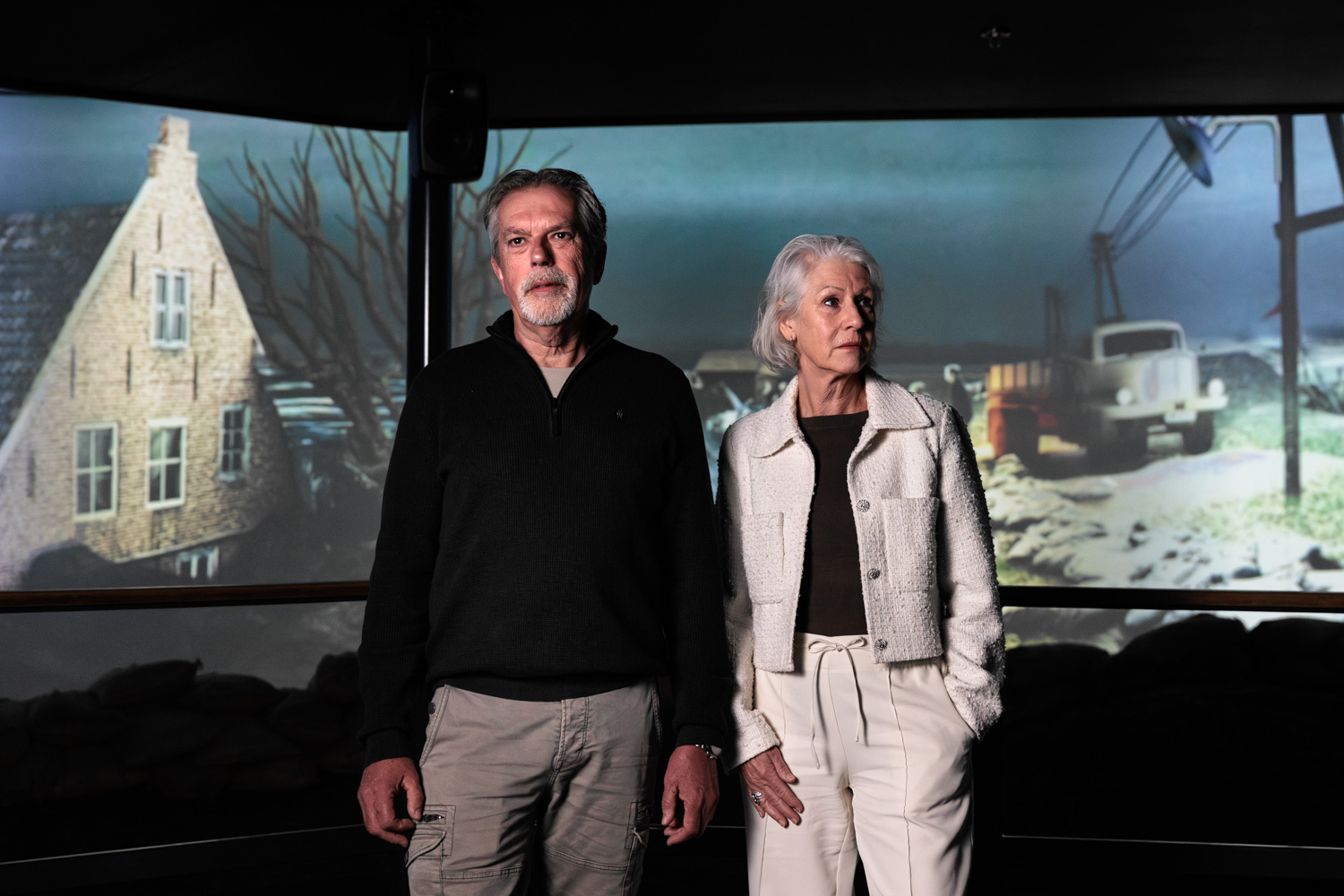 Oudere man en vrouw staan in een tentoonstelling over de Watersnoodramp van 1953, met projectie van een overstroomd dorp op de achtergrond.
