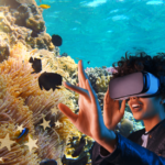 Persoon met een VR-bril die enthousiast haar hand uitstrekt, terwijl ze een virtuele onderwaterwereld met koraalriffen en kleurrijke vissen ervaart.