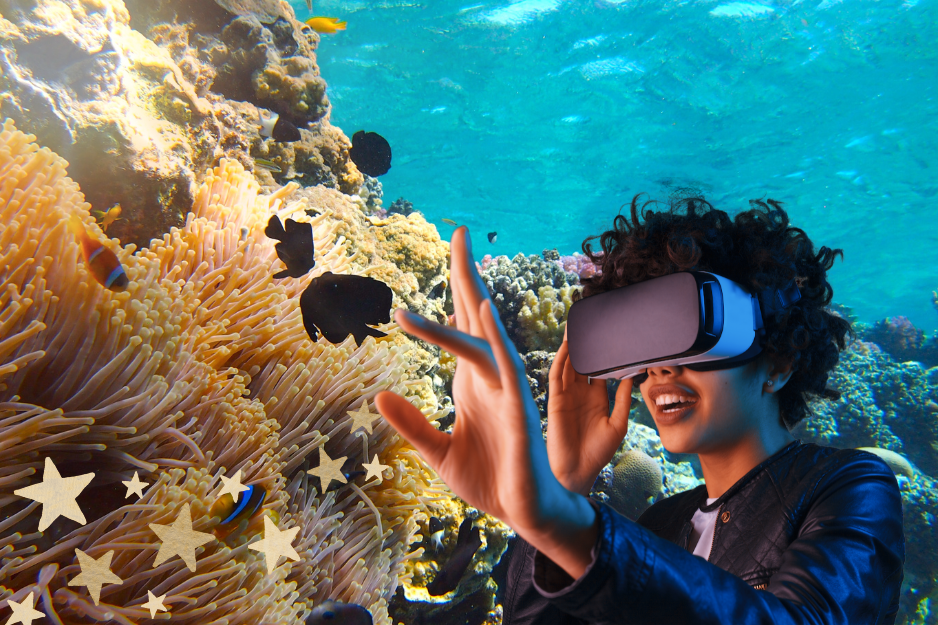 Persoon met een VR-bril die enthousiast haar hand uitstrekt, terwijl ze een virtuele onderwaterwereld met koraalriffen en kleurrijke vissen ervaart.